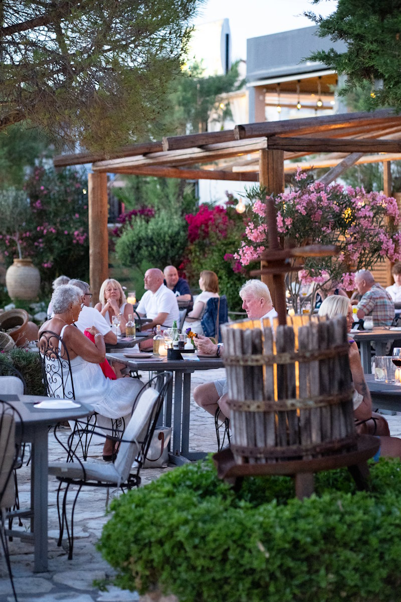 Guests dining at a full outdoor garden at Zakanthi Restaurant at dusk, with bougainvillea, oleander and wooden pergola, Kalamaki Zakynthos